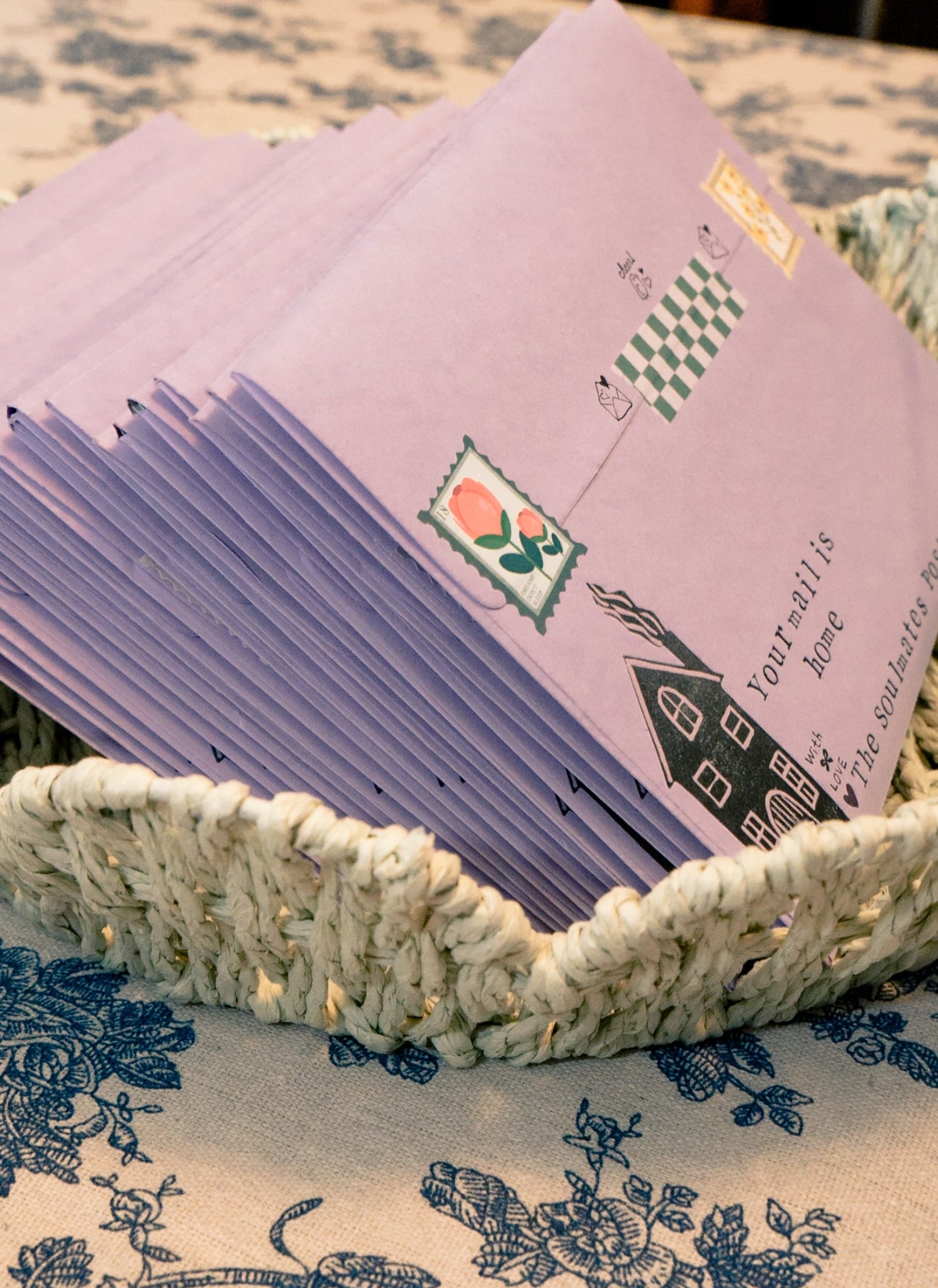 Stack of purple greeting cards with green and red designs in a woven basket on a floral tablecloth.